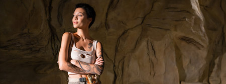 tattooed archaeologist in sexy tank top standing with crossed arms and looking away in cave, bannerの写真素材