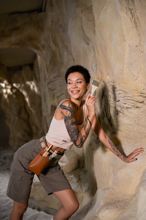 smiling tattooed archaeologist in tank top looking away while working with brush near rock in caveの写真素材