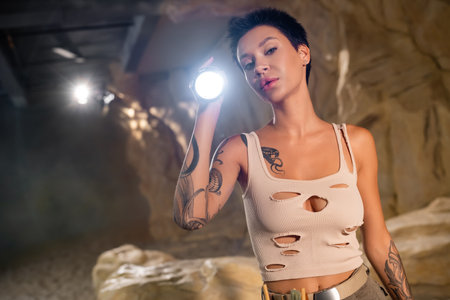young tattooed archaeologist in tank top holding flashlight and looking at camera in caveの写真素材