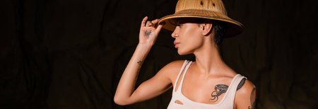 young tattooed archaeologist in tank top adjusting safari hat in dark desert, bannerの写真素材