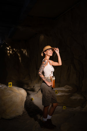 sexy tattooed archaeologist adjusting safari hat and smiling at camera near numbered cards in dark caveの写真素材