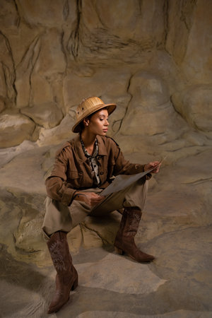 archaeologist in safari hat and cowboy boots looking at ancient map in cave