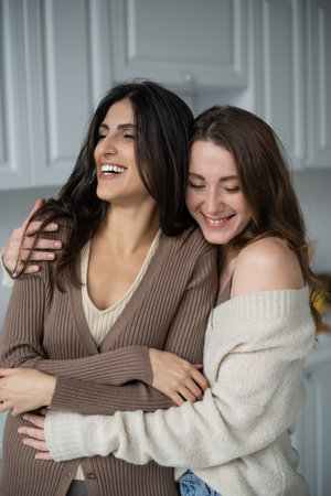 Cheerful woman in sweater hugging girlfriend at homeの写真素材