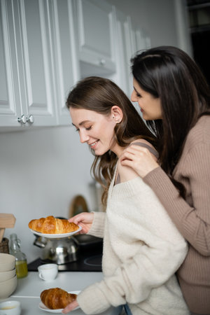 Young woman hugging partner in cozy sweater holding croissants in kitchenの写真素材