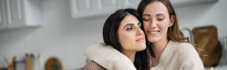 Positive woman in sweater hugging girlfriend in kitchen, bannerの写真素材