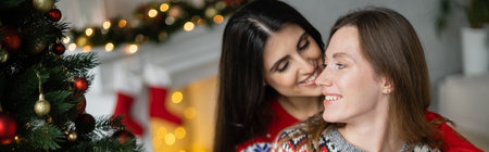 Smiling woman kissing girlfriend in sweater near christmas tree at home, bannerの写真素材