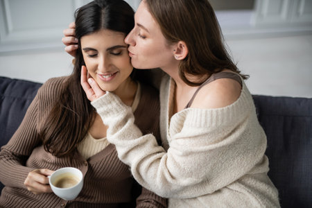 Lesbian woman in warm sweater kissing girlfriend with cup of coffee on couchの写真素材