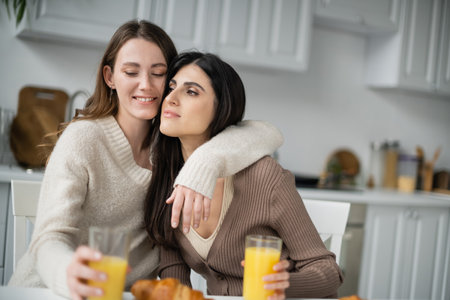 Young lesbian woman hugging girlfriend near orange juice and croissant in kitchenの写真素材