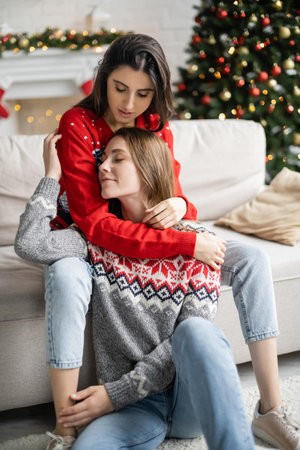 Young woman in sweater hugging girlfriend during christmas celebration at homeの写真素材
