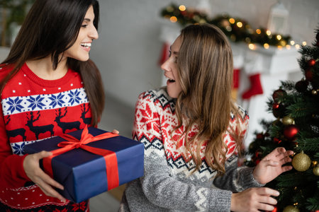 Woman in sweater holding christmas gift near amazed girlfriend in living roomの写真素材