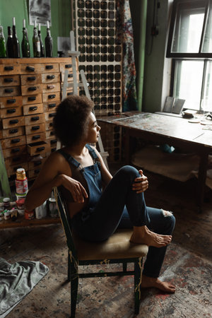 pretty and dreamy african american artist in overalls sitting barefoot in workshop and looking away through windowの写真素材