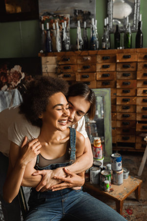 young artist embracing cheerful african american woman near vintage furniture in workshopの写真素材