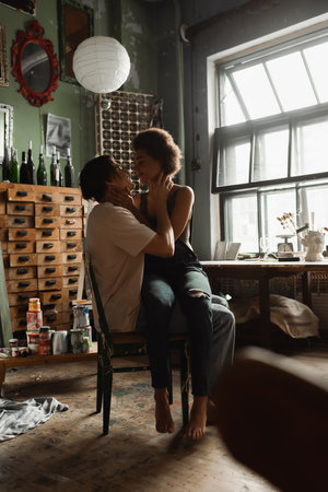 sexy african american woman in overalls sitting on laps of boyfriend in art studio with vintage furniture and large windowの写真素材