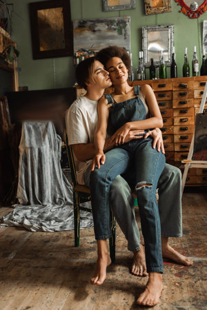 young artist hugging sensual african american woman in denim overalls sitting on his laps with closed eyesの写真素材