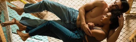 shirtless man in jeans and african american woman in overalls hugging in hammock while lying with...