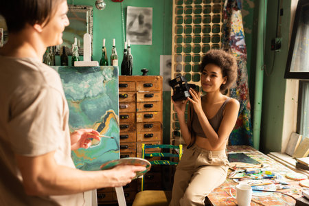 smiling african american woman holding vintage camera near boyfriend standing with palette near...