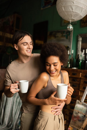 happy man with cup of tea hugging cheerful girlfriend in art workshopの写真素材