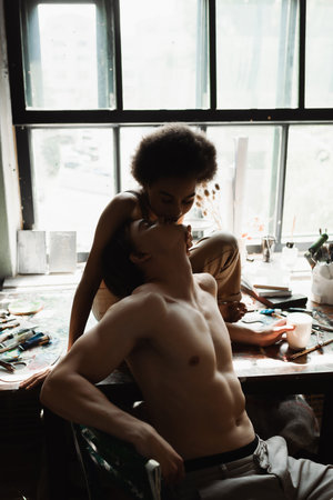 sensual african american woman sitting on windowsill in workshop and kissing sexy shirtless boyfriendの写真素材