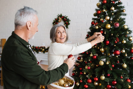 happy middle aged woman with grey hair decorating christmas tree and looking at husband holding wicker basketの写真素材