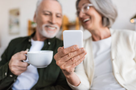 cheerful and mature woman holding smartphone near happy husband with cup of teaの写真素材