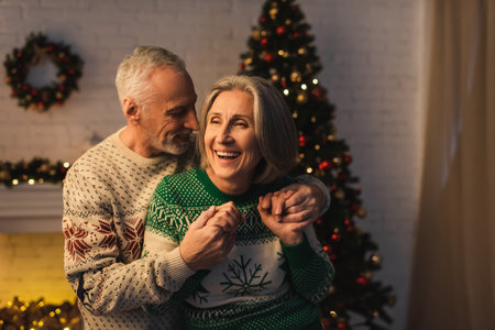 joyful bearded man in festive sweater hugging pleased mature wife near christmas tree in eveningの写真素材