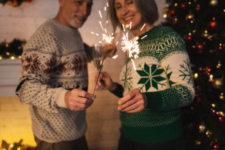 cheerful middle aged couple in festive sweaters holding bright sparklers near blurred christmas treeの写真素材