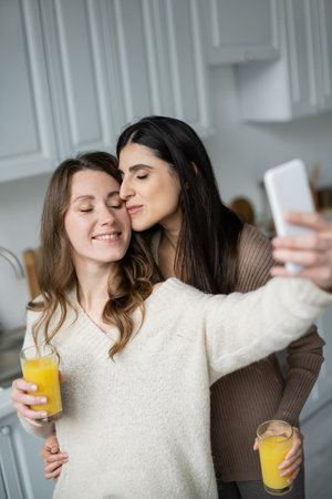 Lesbian woman holding orange juice and kissing girlfriend while taking selfie in kitchenの写真素材