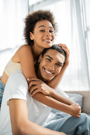 curly african american woman embracing happy boyfriend and looking at camera