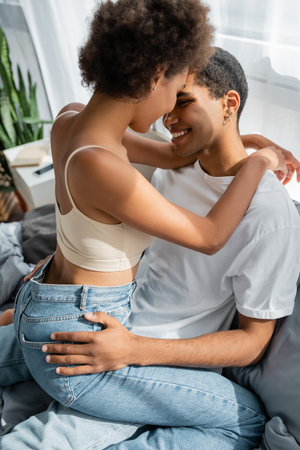 smiling african american woman in jeans near boyfriend embracing her buttocks on bedの写真素材