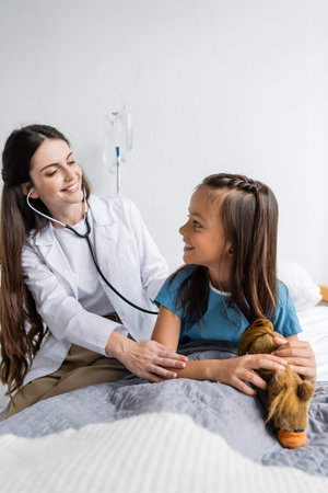 Cheerful pediatrician with stethoscope calming patient near soft toy on hospital bedの写真素材
