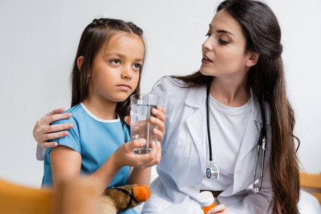 Doctor holding pills and talking to girl with glass of water in hospital wardの写真素材