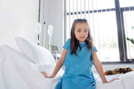 Child in patient gown looking at camera while sitting on bed in hospitalの写真素材