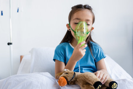 Sick child using oxygen mask near toy on bed in hospitalの写真素材