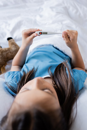 Kid holding electronic thermometer while lying on bed in hospital wardの写真素材