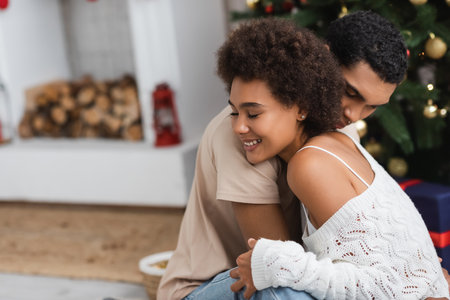 smiling african american woman in white openwork sweater hugging boyfriend on christmas day at homeの写真素材