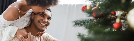 young african american woman embracing and kissing boyfriend near blurred christmas tree, bannerの写真素材