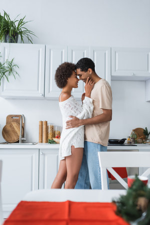 side view of african american woman in white sweater embracing with boyfriend in kitchenの写真素材