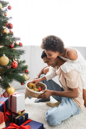 happy african american couple holding wicker basket with baubles near gift boxes and christmas treeの写真素材