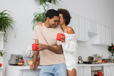 happy african american woman in white sweater holding red cup and kissing man in kitchenの写真素材