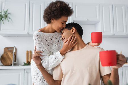 pleased african american couple with red cups hugging in kitchenの写真素材