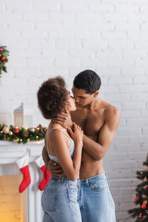 shirtless african american man in jeans embracing passionate girlfriend near decorated fireplaceの写真素材
