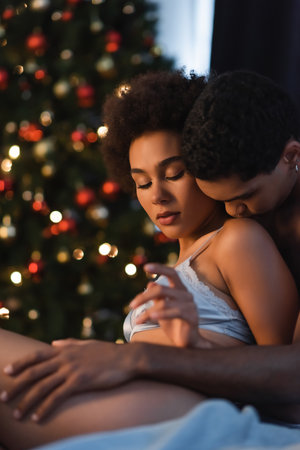 young african american man hugging and kissing sexy girlfriend in bedroom with christmas lightsの写真素材