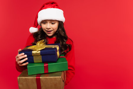 happy girl in santa hat and sweater holding bunch of Christmas presents isolated on redの写真素材