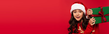 amazed girl in santa hat and sweater holding wrapped Christmas gift box isolated on red, bannerの写真素材