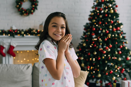 Happy girl in pajama looking at camera and doing praying hands during Christmas celebration at homeの写真素材