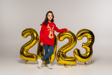 full length of cheerful child in red sweater standing near balloons with 2023 numbers on greyの写真素材