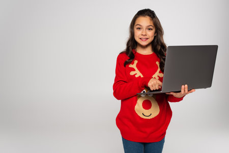 smiling child in red Christmas sweater using laptop isolated on greyの写真素材
