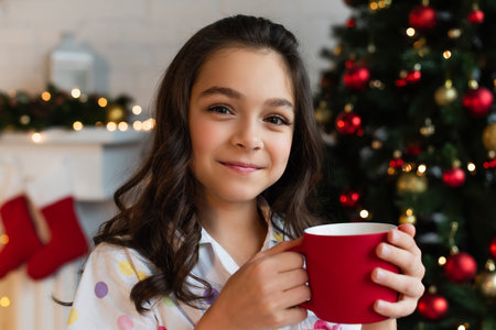 Preteen girl in pajama holding cup and looking at camera near blurred Christmas tree at home in eveningの写真素材