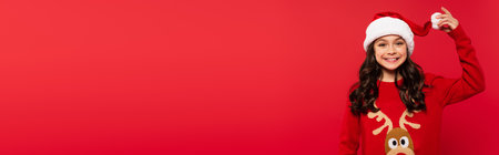 happy girl in sweater holding pompon on santa hat and smiling isolated on red, bannerの写真素材