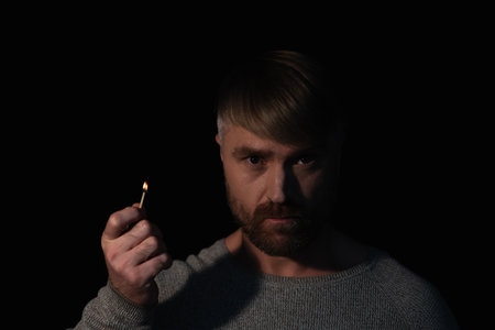 man in darkness holding lit match and looking at camera during electricity blackout isolated on blackの写真素材